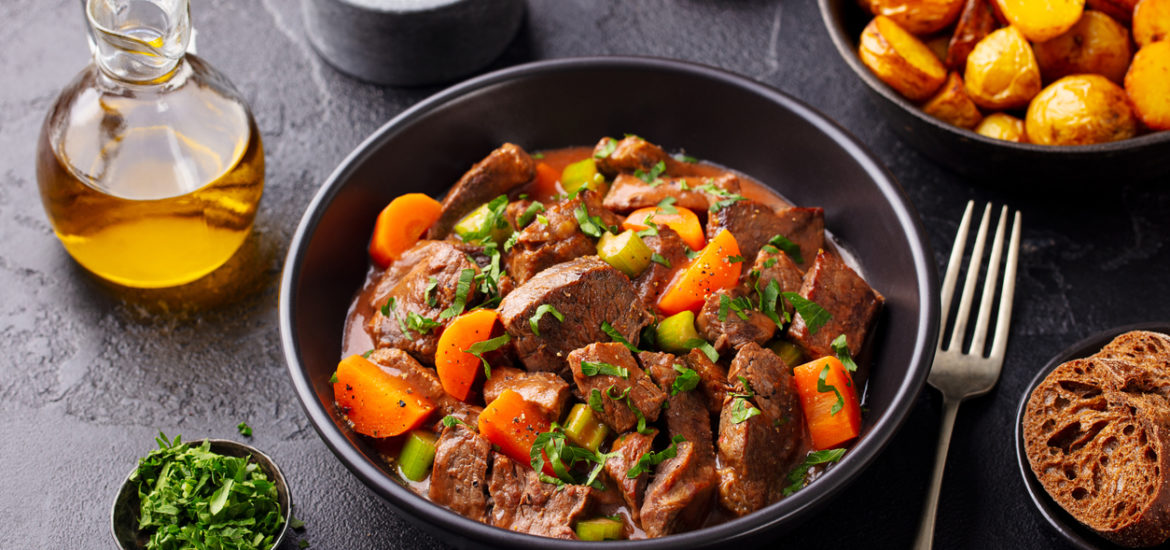Beef meat and vegetables stew in black bowl with roasted baby potatoes. Dark background.