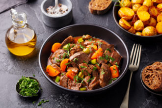 Beef meat and vegetables stew in black bowl with roasted baby potatoes. Dark background.